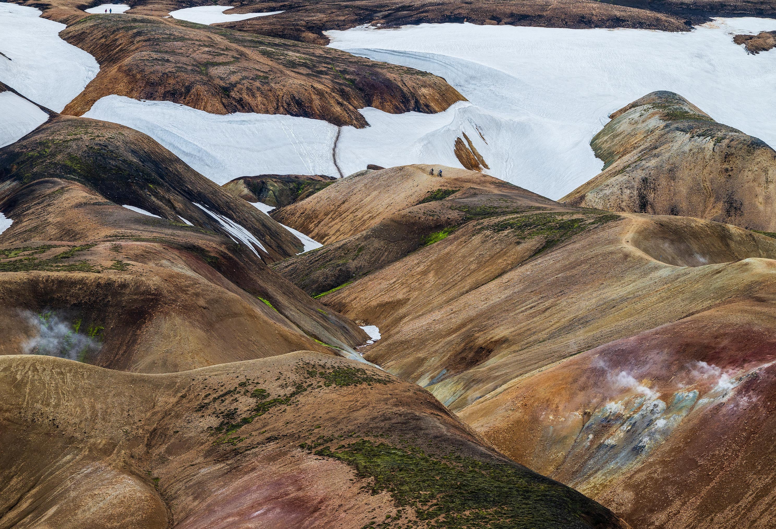 Colorful landscape with brown, green, and white hues in Iceland with bikers on a ridge