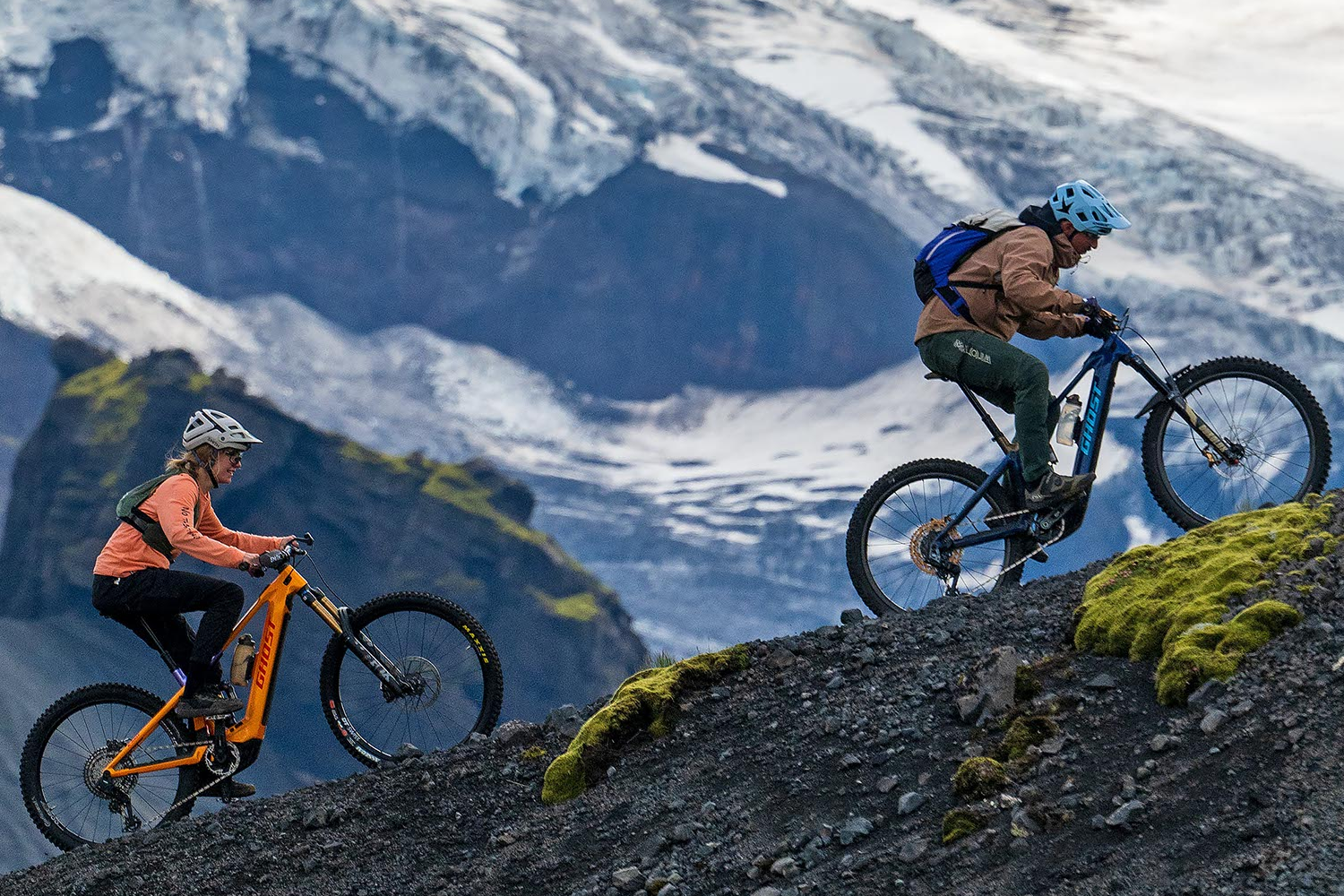 Two mountain bikers on a trail with snow-capped mountains in the background