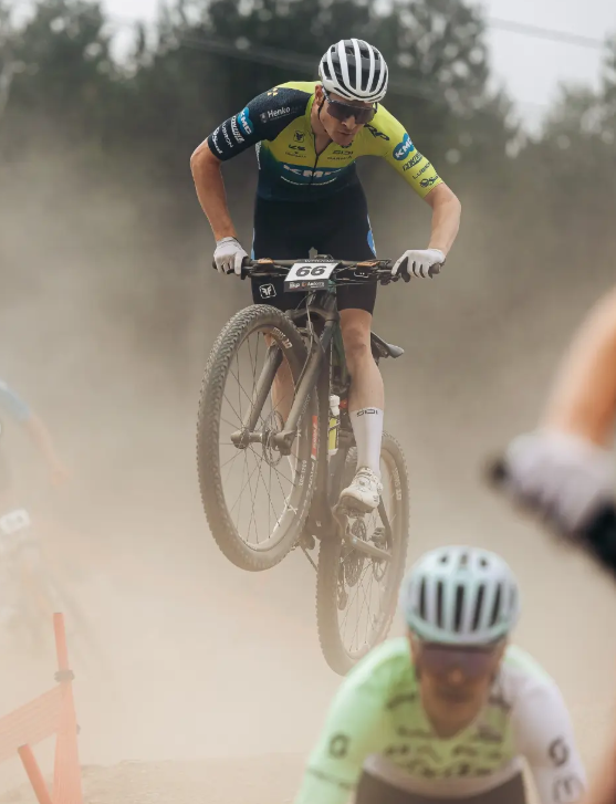 A mountain biker mid-jump during a dusty outdoor race, wearing a yellow and blue cycling jersey, black shorts, white gloves, and a white helmet. Another cyclist is blurred in the foreground.