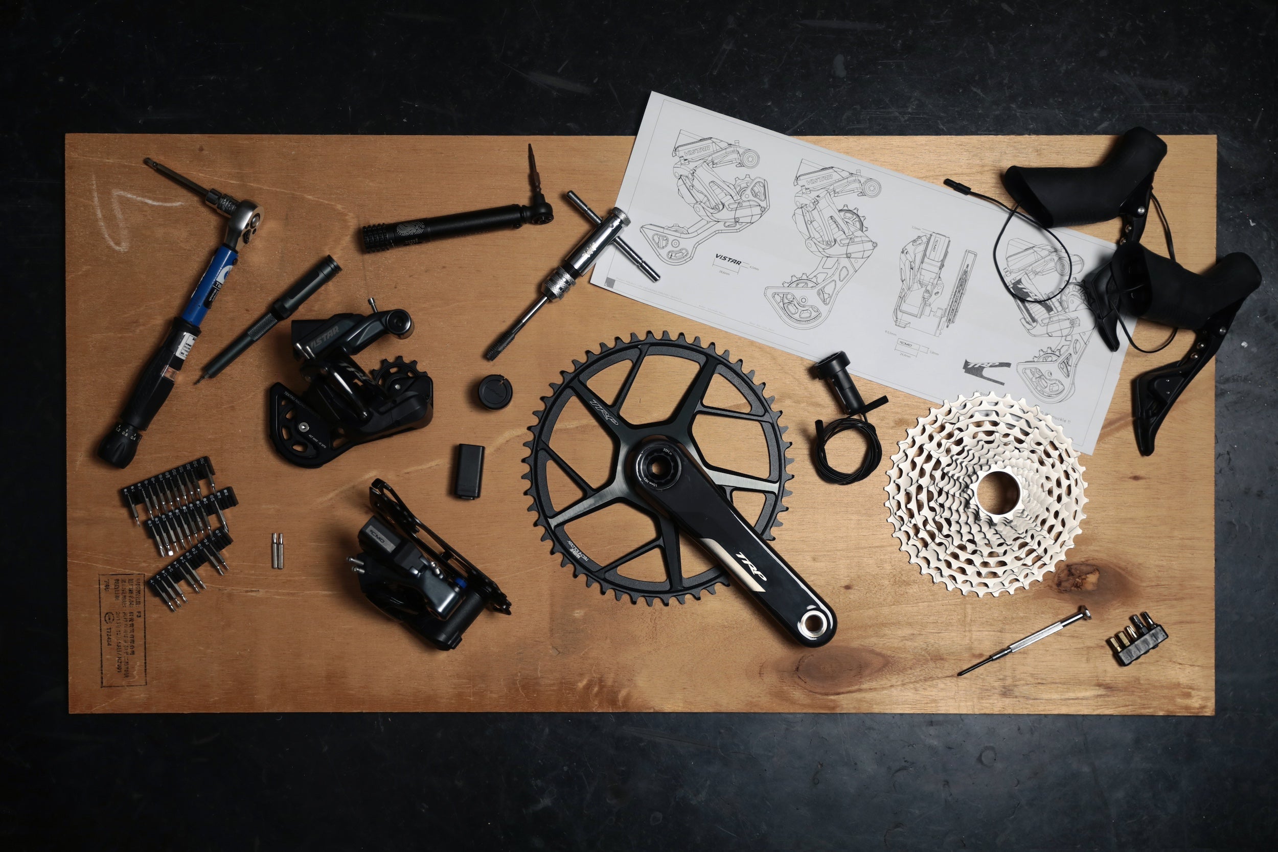 assortment of TRP VISTA components for road and gravel bikes like derailleur, chainring with crank and shifters on a table with tools and  technical drawings