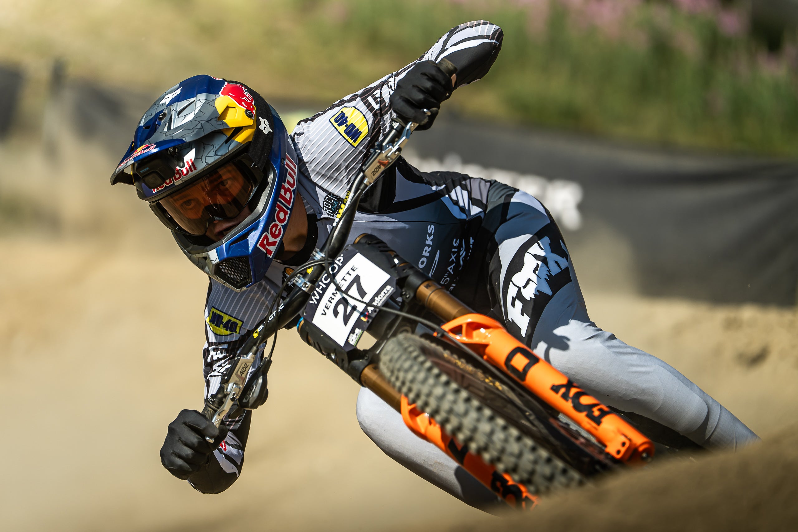 A female bike racer leans sharply into a turn on an orange and black Fox dirt bike, wearing a blue, white, and black Red Bull helmet and matching sponsor apparel on a dirt track.