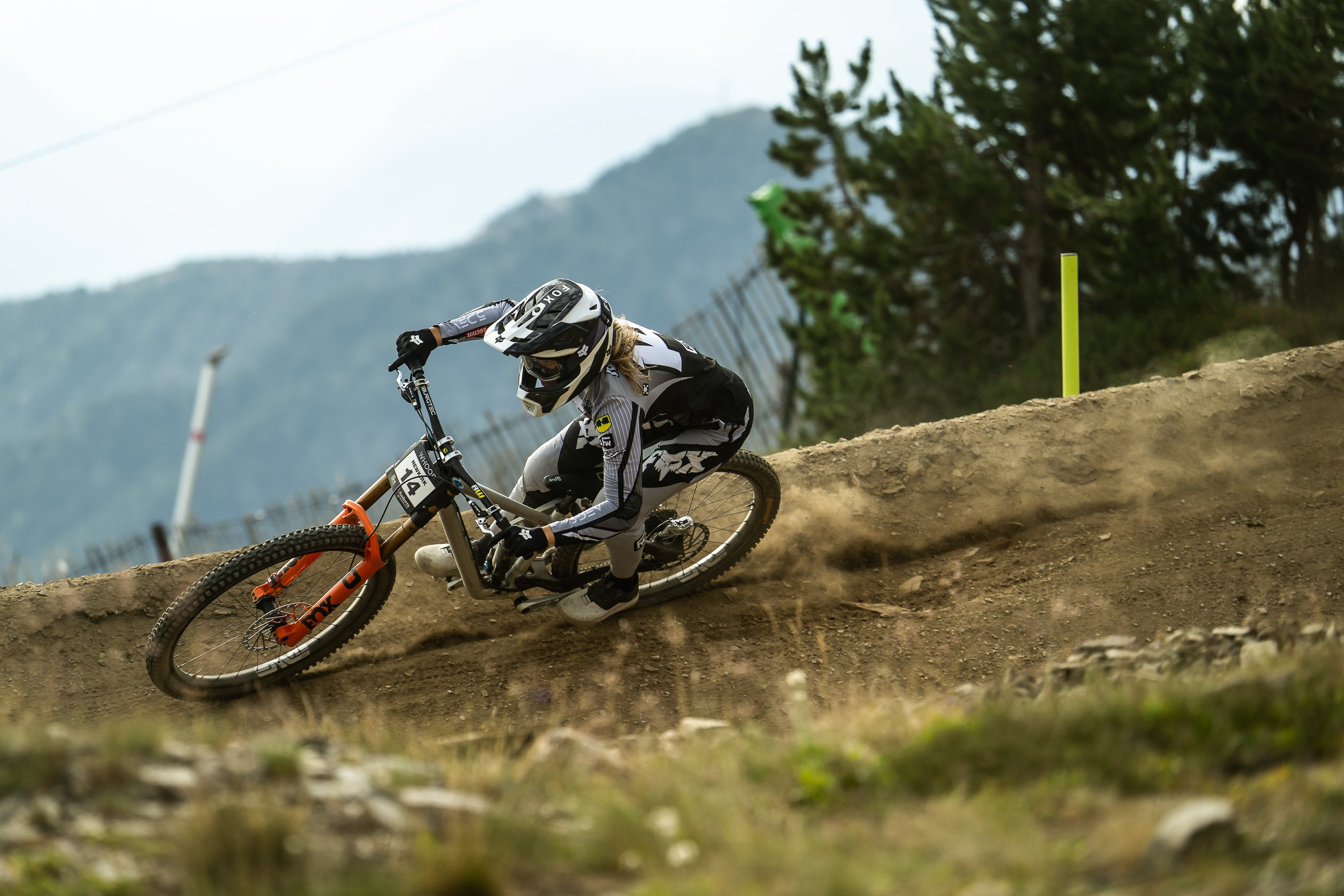 A female mountain biker in full racing gear leans sharply into a turn on a dirt track, with trees and mountains blurred in the background.