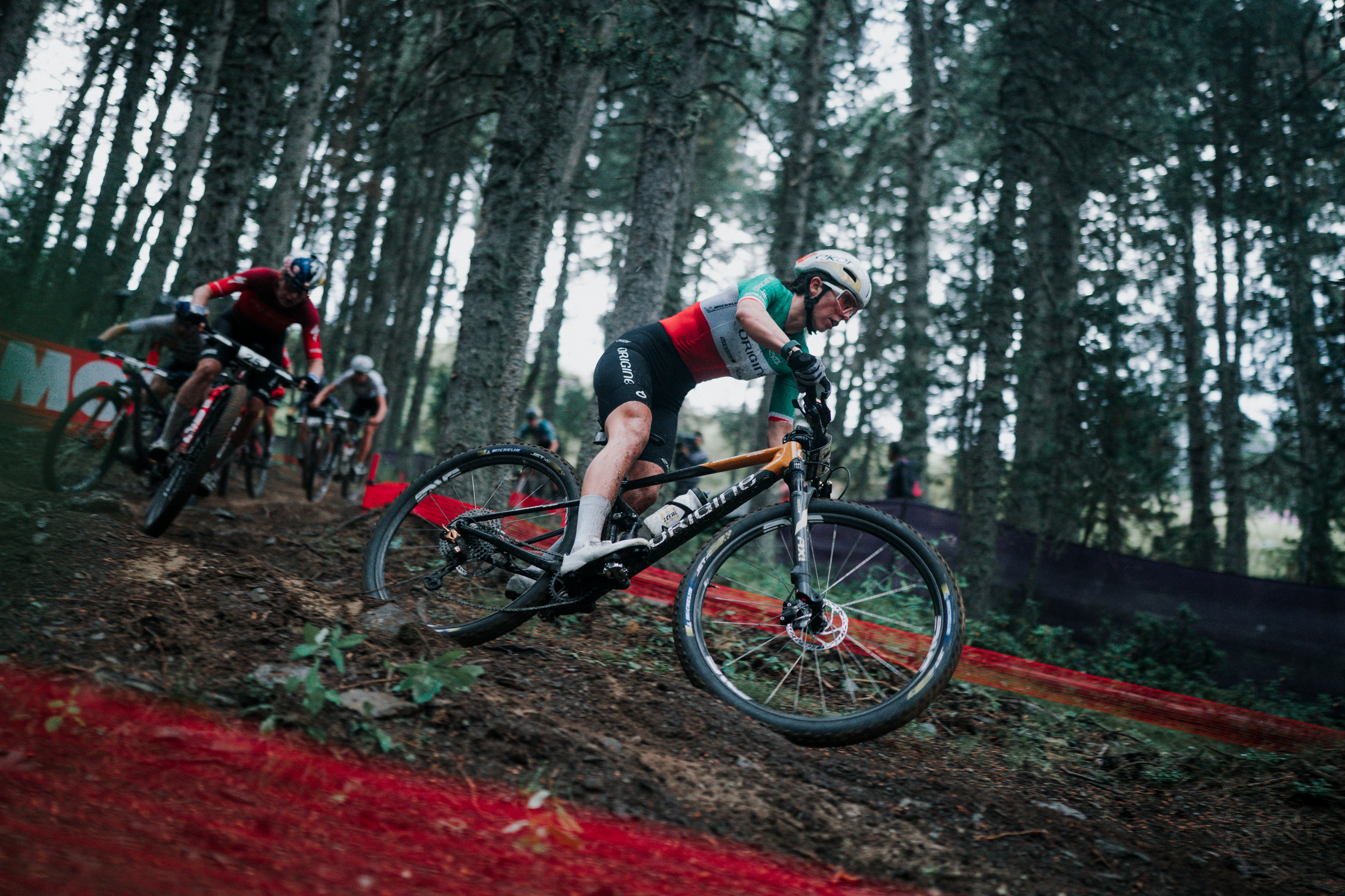 A female mountain biker in a forest course wearing a red, black, and green jersey, riding a black and orange Santa Cruz mountain bike on a dirt track with other racers in the background.